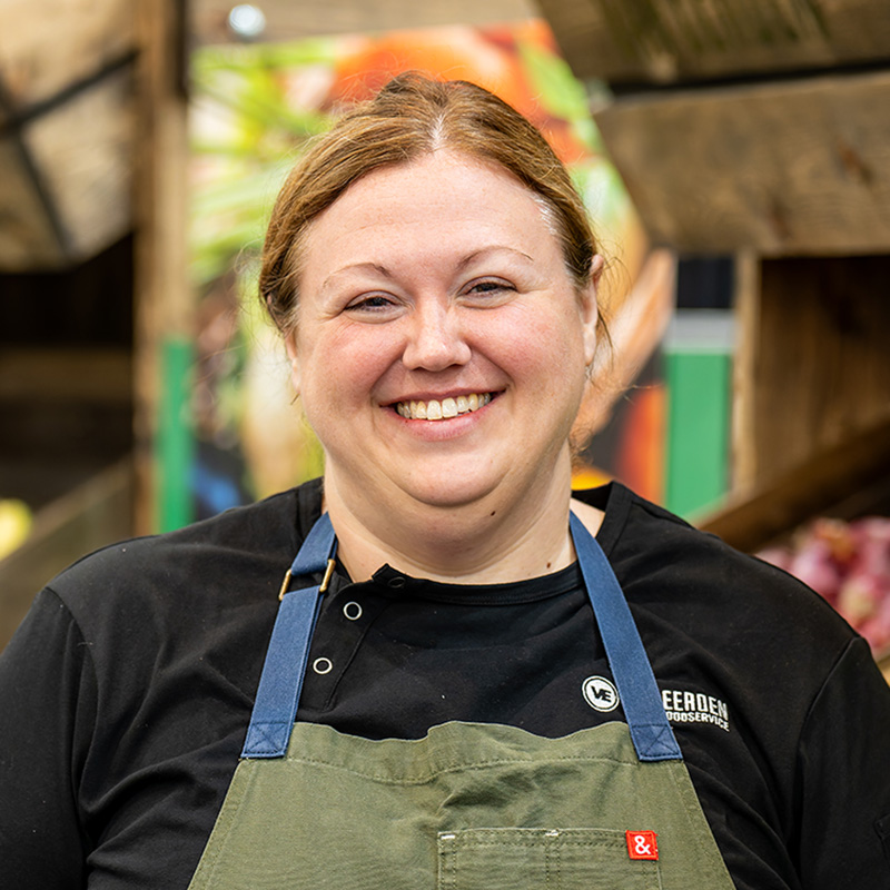 Cheerful chef in green apron smiles warmly at vibrant market, showcasing her love for food.