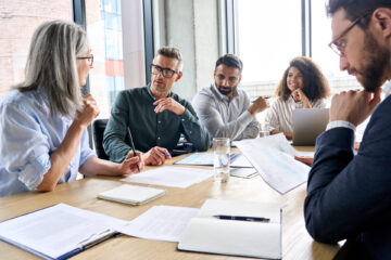Professional diverse team engaged in collaborative business meeting at wooden table with laptops and documents in bright office setting.