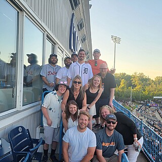 A group of employees enjoying a corporate team-building outing at a baseball stadium, gathered on an upper deck with stadium field and seating visible below.
