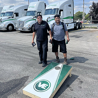 Two professional staff members playing cornhole in front of green and white refrigerated food distribution trucks, representing logistics and supply chain partnerships.