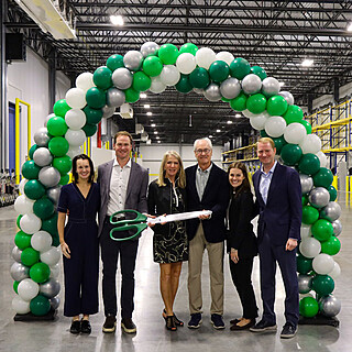 Executive team conducting ribbon cutting ceremony at food distribution warehouse facility with green and white balloon arch decoration.