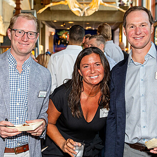 Three business professionals smiling at a food service industry networking event, holding appetizers and name badges.