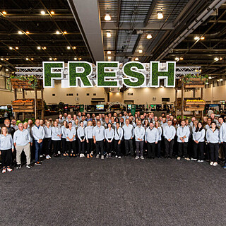 Large team of food distribution professionals in white shirts posing together at a wholesale food facility under a FRESH sign, representing B2B foodservice sales and supply chain excellence.