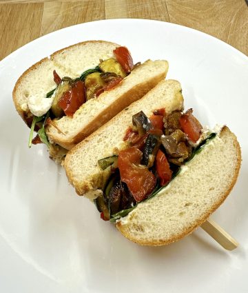 Colorful grilled vegetable sandwich with creamy spread, fresh greens, and toasted bun on a white plate.