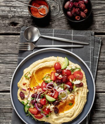 Refreshing Greek hummus topped with veggies, olives, and feta on a rustic wooden table.