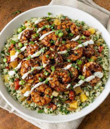 Glazed shrimp rice bowl with herbs, vegetables, and creamy drizzle on rustic wooden background.