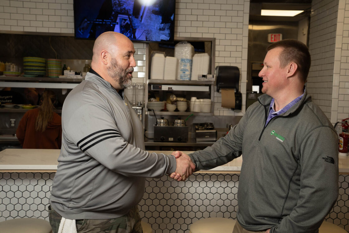 Two professionals shake hands in a commercial kitchen setting, representing a B2B food service or wholesale food distribution business agreement.