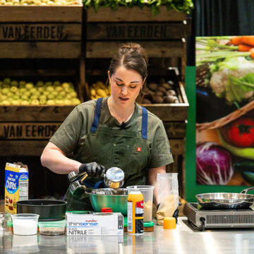Professional chef in apron preparing fresh vegetables in a commercial foodservice kitchen with wholesale produce crates and supplies visible.