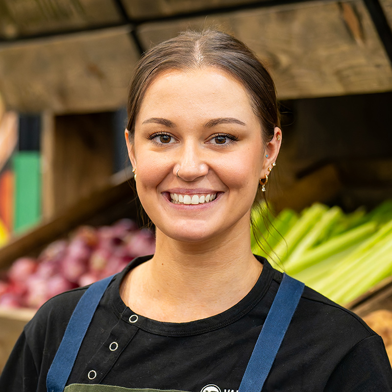 Cheerful vendor at a vibrant market, showcasing fresh produce and inviting customers to explore.
